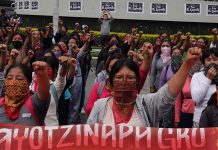 Multiple women standing in rows in a courtyard with their left fists raised in the air. Before the front row is a banner saying "Ayotzinapa, Guerrero."