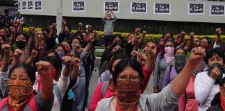 Multiple women standing in rows in a courtyard with their left fists raised in the air. Before the front row is a banner saying "Ayotzinapa, Guerrero."