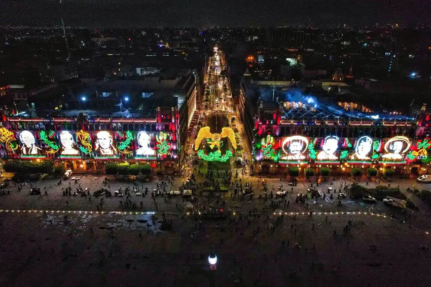 Drone shot of Mexico City's main square, the Zocalo, lit up with a row of images of Mexican independence and revolutionary figures' faces projected on the Zocalo's buildings, with a lit image of Mexico's national symbol, an eagle with its wings spread displayed over the road into the Zocalo