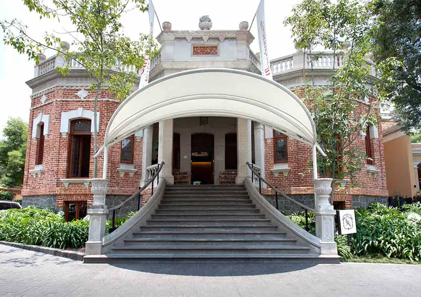 The El Cardenal Restaurant in the San Angel neighborhood of Mexico City. The building is a historic brick mansion with a wide marble staircase, three rooftop turrets and an expansive canopy that covers the base of the staircase.