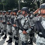 Horizontal line of Mexico's national guard members in uniform and holding guns, saluting ceremonially