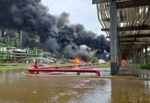 Black smoke pouring out of a refinery in the background, with flames at the base. In the foreground is a river with red pipes spewing what looks like water.