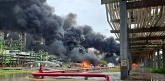 Black smoke pouring out of a refinery in the background, with flames at the base. In the foreground is a river with red pipes spewing what looks like water.