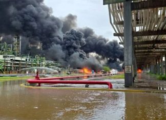 Black smoke pouring out of a refinery in the background, with flames at the base. In the foreground is a river with red pipes spewing what looks like water.