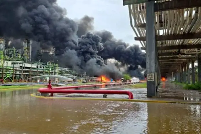 Black smoke pouring out of a refinery in the background, with flames at the base. In the foreground is a river with red pipes spewing what looks like water.