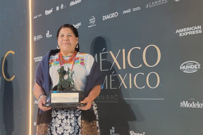 Restauranteur Rosalba Morales Bartolo poses against a backdrop of the Canirac Awards, poses for photos with her trophy. She's wearing a white, traditional indigenous blouse with handmade embroidery and a dark blue shawl.