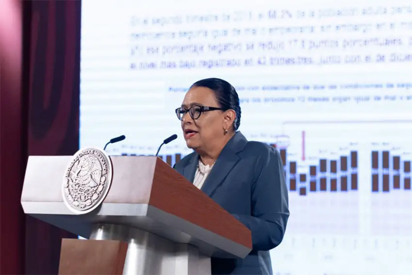 Rosa Icela Rodríguez de pie en un podio en una conferencia de prensa hablando con la prensa con un informe proyectado en una pantalla detrás de ella.