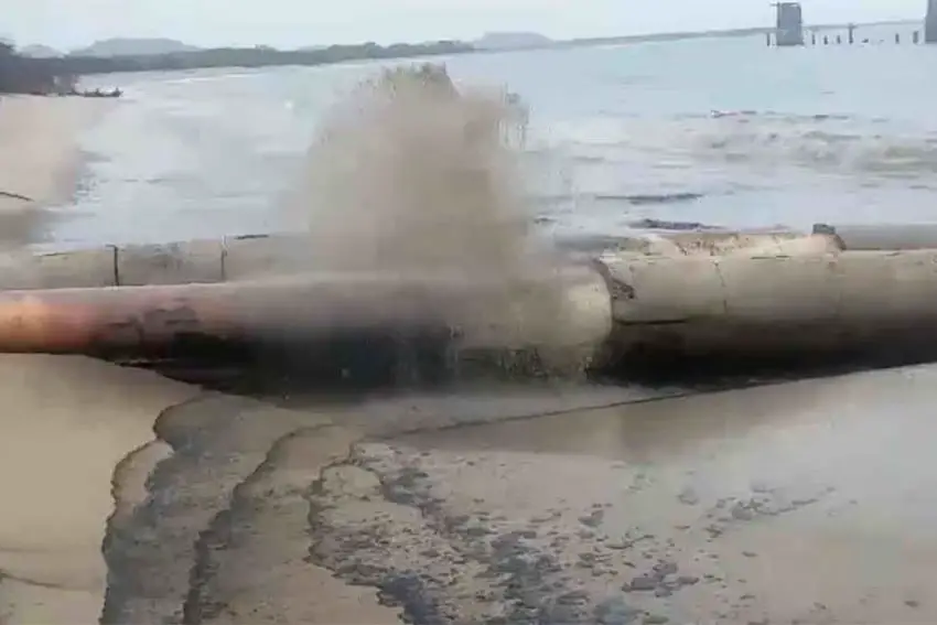 Two pipelines side by side on the shore of a river that feed into a river. They are emitting gray smoke.