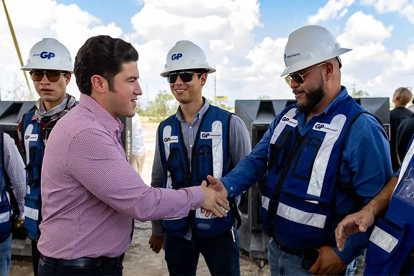 Gov. Garcia in a pink shirt shaking hands with a male worker who is in jeans, a hardhat, and a uniform shirt bearing the logo for the company GP. Two other male workers in the same type of outfits look on.