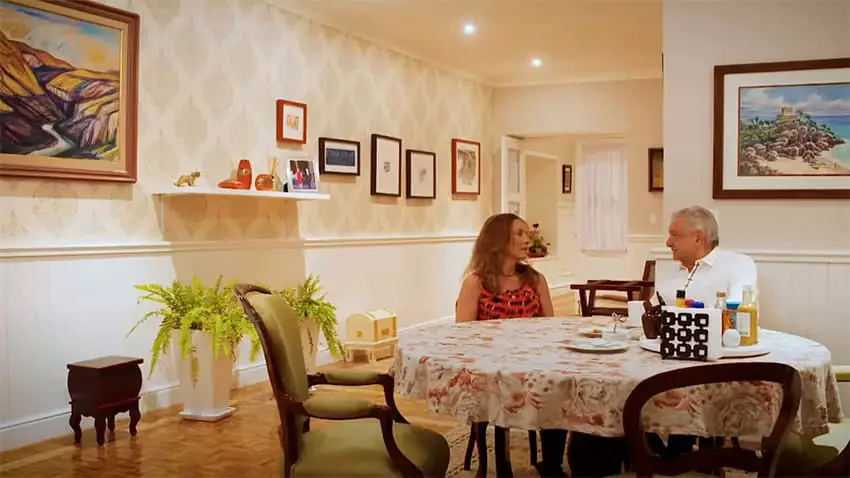 An elderly couple sit in a traditional, brightly lit dining room with pictures on the walls