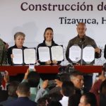Mexico's President Sheinbaum and members of her cabinet standing in front of a finished wooden table holding up government paperwork related to the Mexico City-Pachucha passenger train inside portfolio booklets.