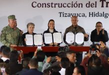 Mexico's President Sheinbaum and members of her cabinet standing in front of a finished wooden table holding up government paperwork related to the Mexico City-Pachucha passenger train inside portfolio booklets.