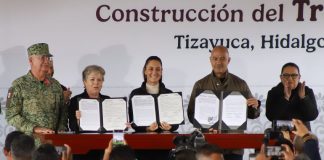 Mexico's President Sheinbaum and members of her cabinet standing in front of a finished wooden table holding up government paperwork related to the Mexico City-Pachucha passenger train inside portfolio booklets.