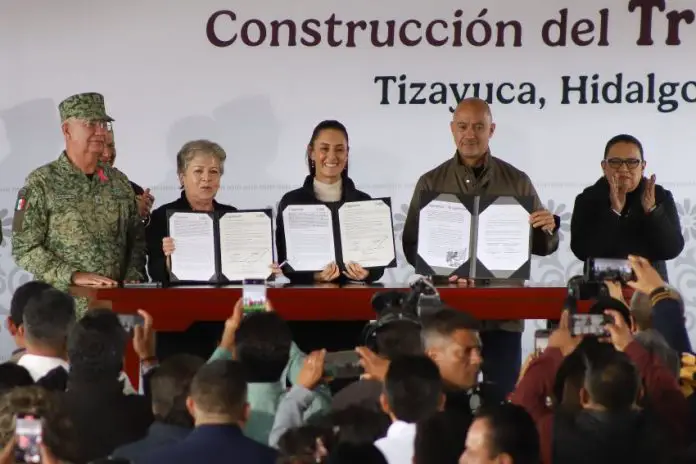 Mexico's President Sheinbaum and members of her cabinet standing in front of a finished wooden table holding up government paperwork related to the Mexico City-Pachucha passenger train inside portfolio booklets.