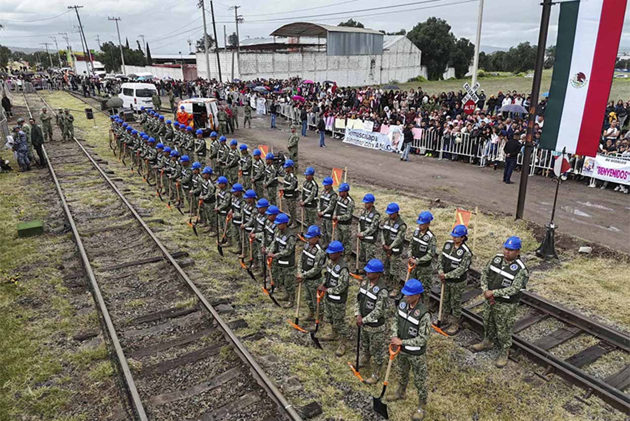 Dos filas horizontales de ingenieros militares con cascos y de pie en posición de firmes, sosteniendo palas frente a ellos en la ubicación del tren interurbano planeado entre Ciudad de México y Pachuca. Cada fila de ingenieros se encuentra frente a un conjunto de vías de ferrocarril.