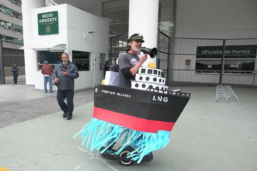 A protester from an environmental organization protesting outside Mexico's Environment Ministry headquarters. He is wearing a captain's hat and has a large paper mache boat tied to his body that makes him look as if he is inside the boat. He is using a megaphone.