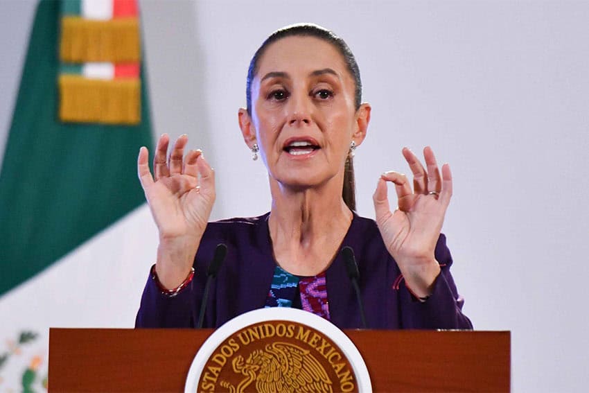 Mexican President Claudia Sheinbaum at a presidential press conference standing behind a podium and holding her hands up near her face, with her thumbs and forefingers of each hand touching. She is in mid-speech.