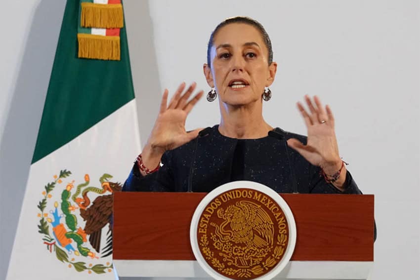 President Claudia Sheinbaum behind a podium at a press conference with her hands raised in front of her shoulders, as if gesturing.