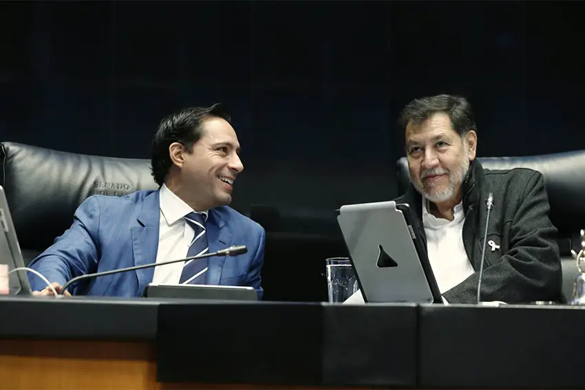 Two Mexican senators sitting at their desks on the Senate floor. One is smiling at looking at the other, who looks straight ahead as if watching proceedings.