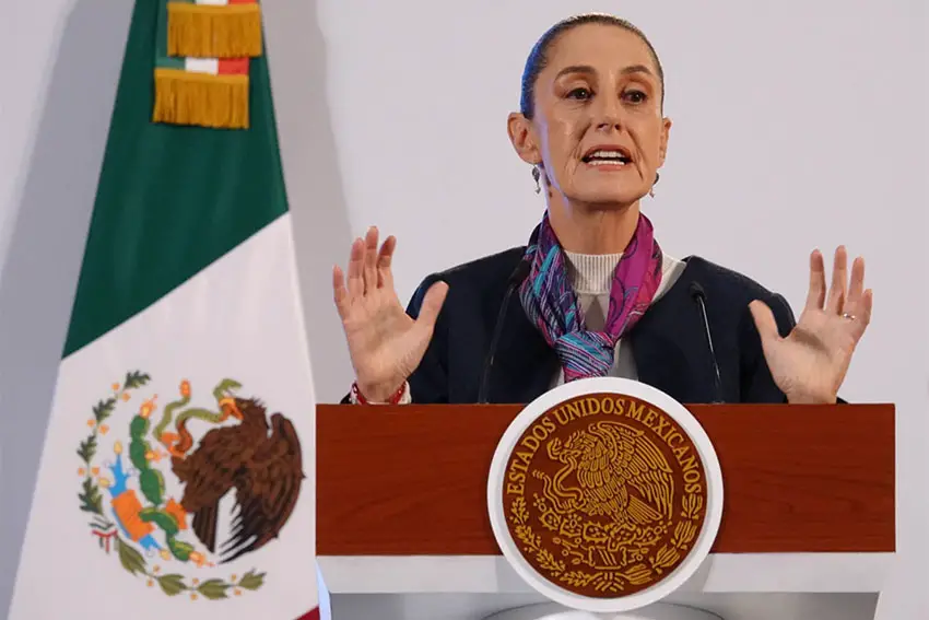 President Sheinbaum standing at a podium at a press conference with her hands in front of her, gesturing as she speaks. Behind her is the Mexican flag.