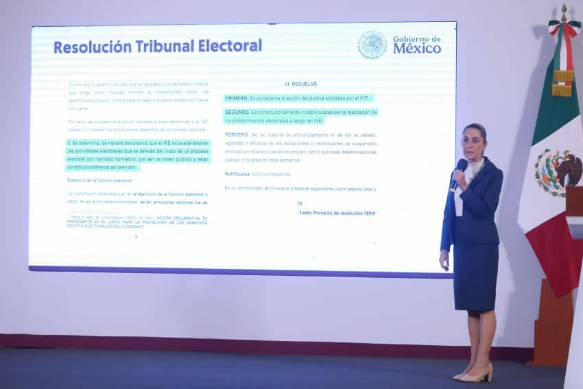 President Sheinbaum speaks into a microphone on a stage in front of a projected slide showing a selection of highlighted text from the Federal Electoral Tribunal ruling on Mexico's 2025 judicial elections