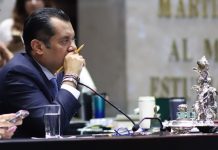 Member of Mexican Chamber of Deputies in a suit at his legislative desk in session. He has a pencil in between his fingers and is holding that hand up to his mouth as he listens.