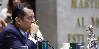 Member of Mexican Chamber of Deputies in a suit at his legislative desk in session. He has a pencil in between his fingers and is holding that hand up to his mouth as he listens.