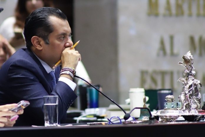 Member of Mexican Chamber of Deputies in a suit at his legislative desk in session. He has a pencil in between his fingers and is holding that hand up to his mouth as he listens.