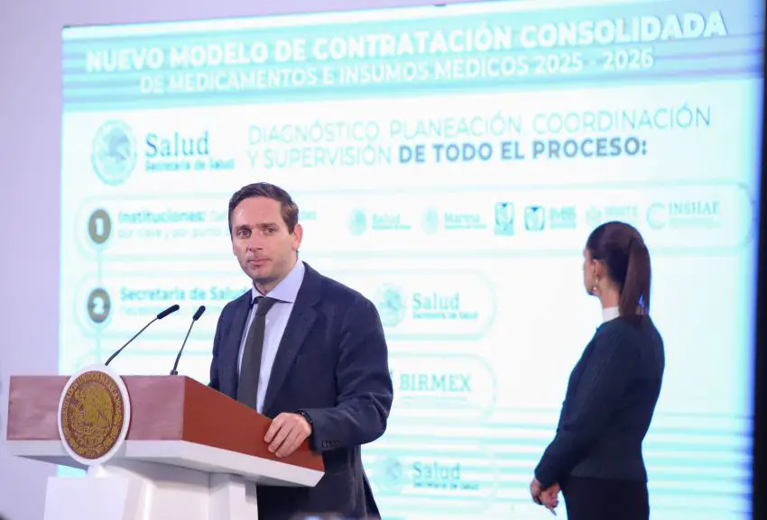 Mexico's Deputy Health Minister Eduardo Clark Garcia at a podium at a press conference, talking to reporters as President Claudia Sheinbaum, behind him, examines a projection screen with his presentation on medications purchasing