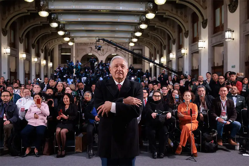 Former Mexico president Lopez Obrador at his last press conference of his presidency standing with his back to the press and his arms crossed in front of him, looking at something off camera. Seated journalists behind him look on.