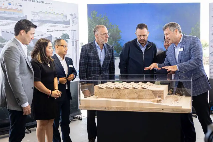 Seis empresarios de pie en un semicírculo mientras una de las personas señala un modelo de un centro de innovación tecnológica de Makino planeado, hecho de madera y colocado detrás de un vidrio.