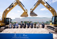 10 people in suits posing for a photo for a photo holding shovels while standing under two bucket loaders whose cranes are bent into something resembling a heart shape.