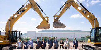 10 people in suits posing for a photo for a photo holding shovels while standing under two bucket loaders whose cranes are bent into something resembling a heart shape.