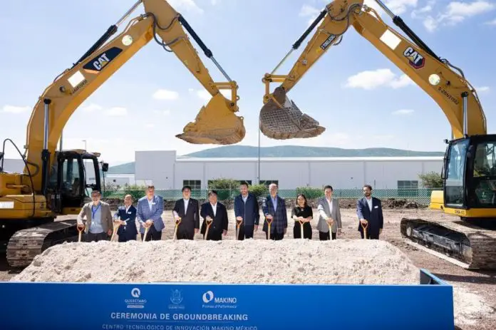 10 people in suits posing for a photo for a photo holding shovels while standing under two bucket loaders whose cranes are bent into something resembling a heart shape.