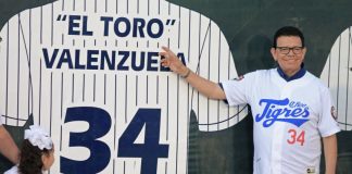 Fernando Valenzuela, wearing a baseball uniform shirt of the Quintana Roo Tigres, stands next to and points at an image on a wall of a larger-than-life version of his baseball shirt for the Los Angeles Dodgers.