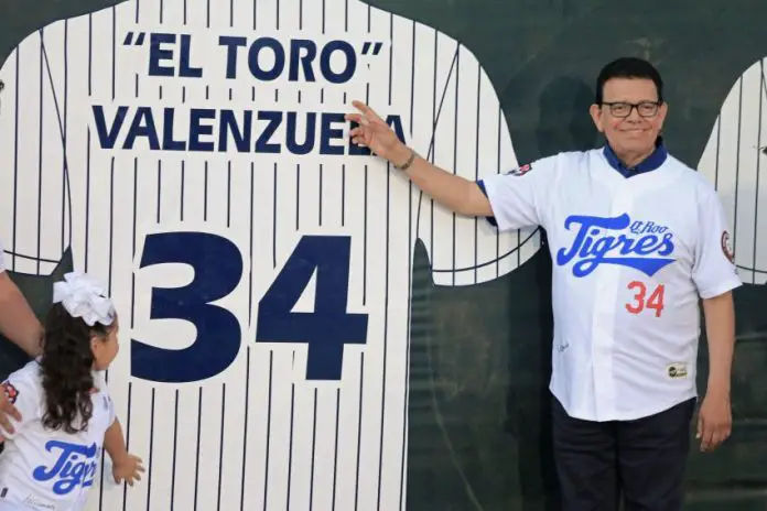 Fernando Valenzuela, wearing a baseball uniform shirt of the Quintana Roo Tigres, stands next to and points at an image on a wall of a larger-than-life version of his baseball shirt for the Los Angeles Dodgers.