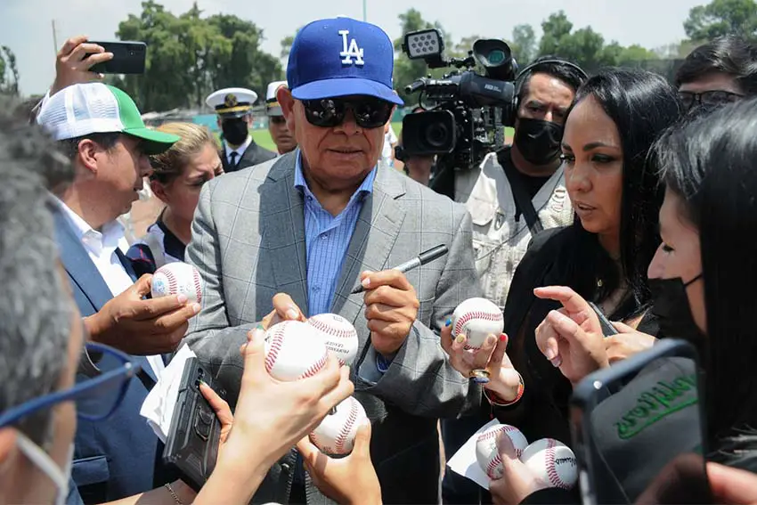 Los Angeles Dodgers baseball legend Fernando Valenzuela signing baseballs held in front of him by people while a man with a news video camera on his shoulder looks on