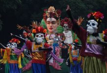 People in a Mexico City Day of the Dead parade from previous years, dressed up in colorful gowns and wearing skeleton masks in the style of Day of the Dead calaveras and standing on a float that features a bust of Frida Kahlo with a crown of flowers.