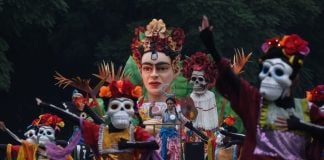 People in a Mexico City Day of the Dead parade from previous years, dressed up in colorful gowns and wearing skeleton masks in the style of Day of the Dead calaveras and standing on a float that features a bust of Frida Kahlo with a crown of flowers.