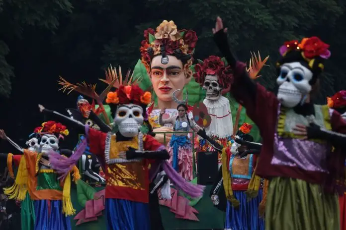 People in a Mexico City Day of the Dead parade from previous years, dressed up in colorful gowns and wearing skeleton masks in the style of Day of the Dead calaveras and standing on a float that features a bust of Frida Kahlo with a crown of flowers.