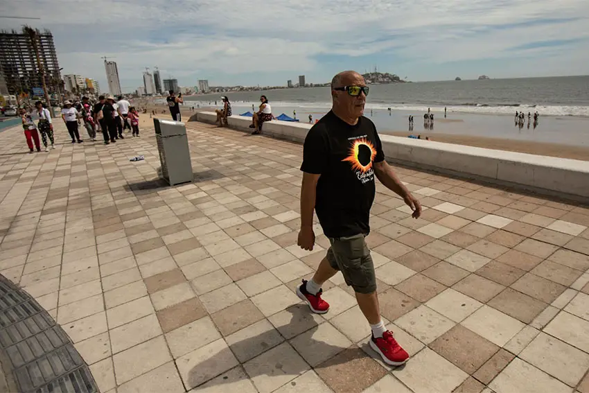 Un hombre con una camiseta con la imagen de un eclipse camina por el malencón bastante vacío de Mazatlán, junto al océano.