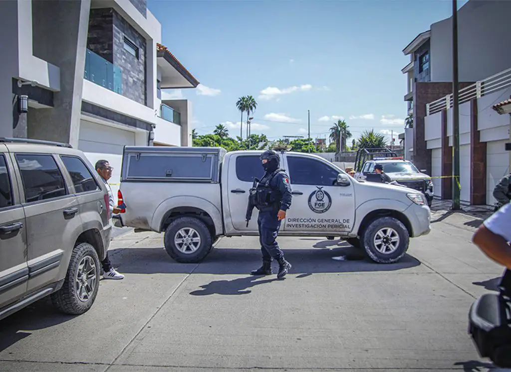 State police officer with a machine gun and wearing a baclava stands at a crime scene where a pickup truck with the Sinaloa attorney general's logo on it is parked, blocking the street horizontally.