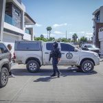 State police officer with a machine gun and wearing a baclava stands at a crime scene where a pickup truck with the Sinaloa attorney general's logo on it is parked, blocking the street horizontally.