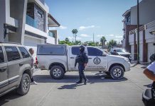 State police officer with a machine gun and wearing a baclava stands at a crime scene where a pickup truck with the Sinaloa attorney general's logo on it is parked, blocking the street horizontally.