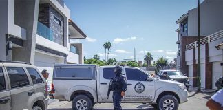 State police officer with a machine gun and wearing a baclava stands at a crime scene where a pickup truck with the Sinaloa attorney general's logo on it is parked, blocking the street horizontally.