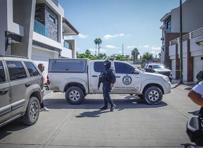 State police officer with a machine gun and wearing a baclava stands at a crime scene where a pickup truck with the Sinaloa attorney general's logo on it is parked, blocking the street horizontally.