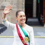 Mexico's President Claudia Sheinbaum smiling and waving at crowds as she stands in front of the National Palace in Mexico City. She is wearing a white dress with traditional embroidery on the cuffs and bearing Mexico's red, white and green presidential sash.