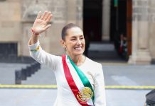 Mexico's President Claudia Sheinbaum smiling and waving at crowds as she stands in front of the National Palace in Mexico City. She is wearing a white dress with traditional embroidery on the cuffs and bearing Mexico's red, white and green presidential sash.