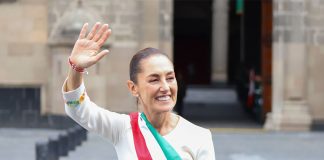 Mexico's President Claudia Sheinbaum smiling and waving at crowds as she stands in front of the National Palace in Mexico City. She is wearing a white dress with traditional embroidery on the cuffs and bearing Mexico's red, white and green presidential sash.
