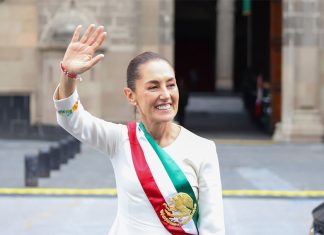Mexico's President Claudia Sheinbaum smiling and waving at crowds as she stands in front of the National Palace in Mexico City. She is wearing a white dress with traditional embroidery on the cuffs and bearing Mexico's red, white and green presidential sash.